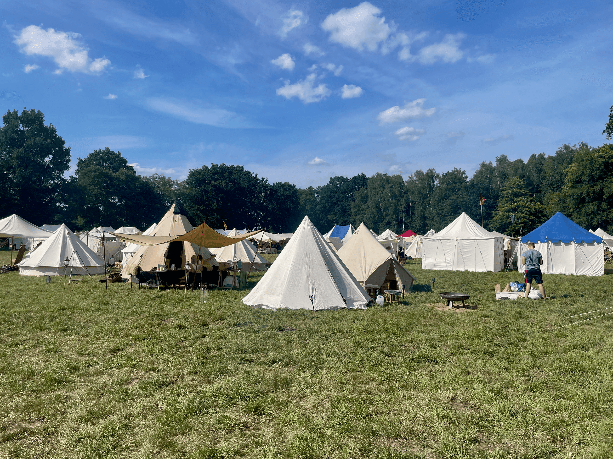 LARP-Lager auf einer Wiese am Waldrand: Reihen heller Leinenzelte und ein beiges Sonnensegel über einer Tischgruppe